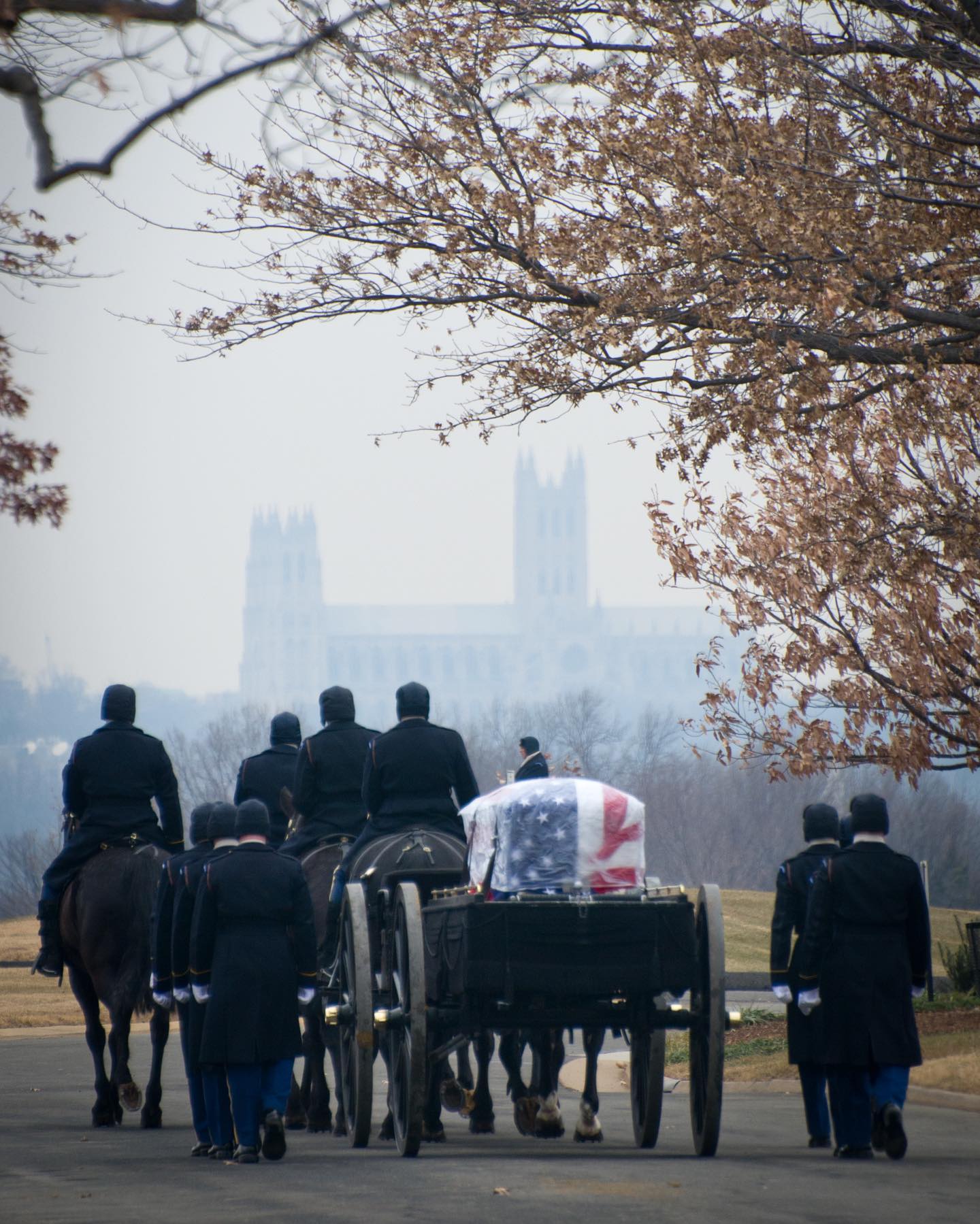 A caisson team can be seen leading a funeral procession through Arlington National Cemetery with the Washington National Cathedral hazy in the background.